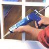 A person uses a blue CRL caulking gun to apply sealant to a window frame.